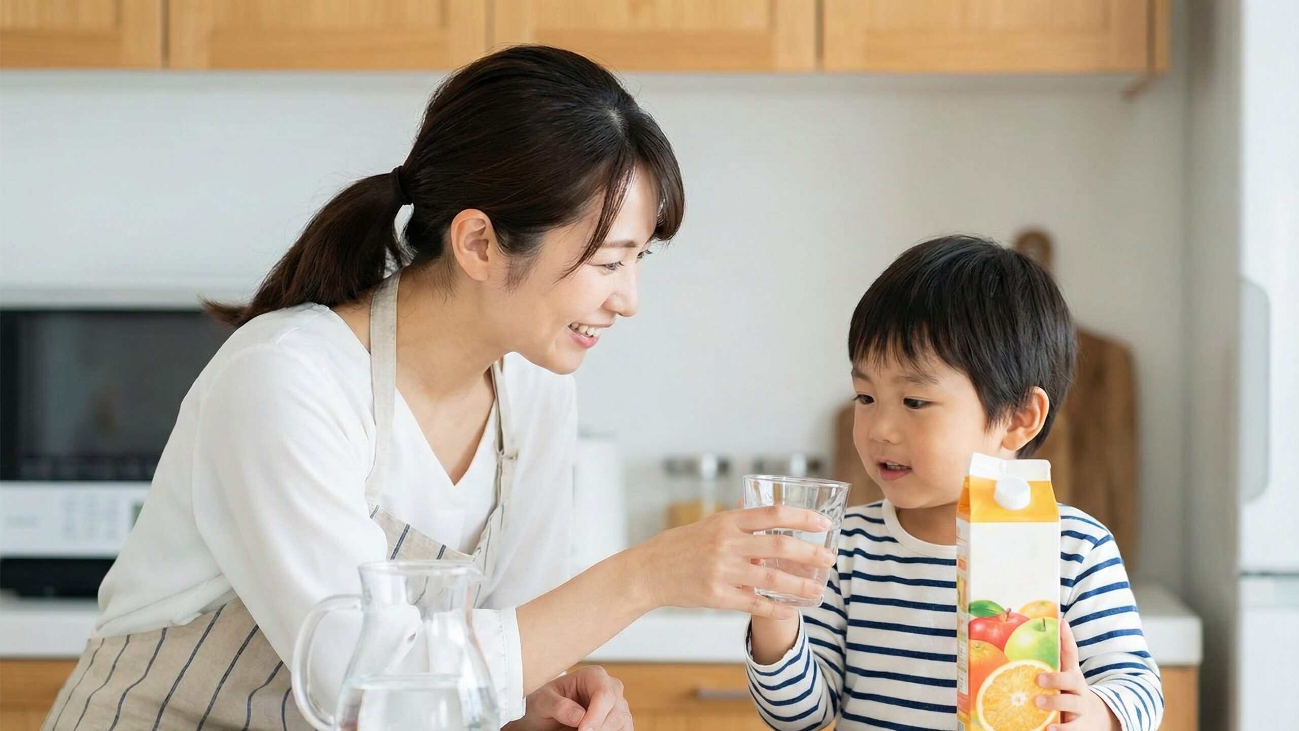 笑顔の母親が、オレンジジュースのパックを持つ男の子に、水が入ったグラスを差し出す場面。