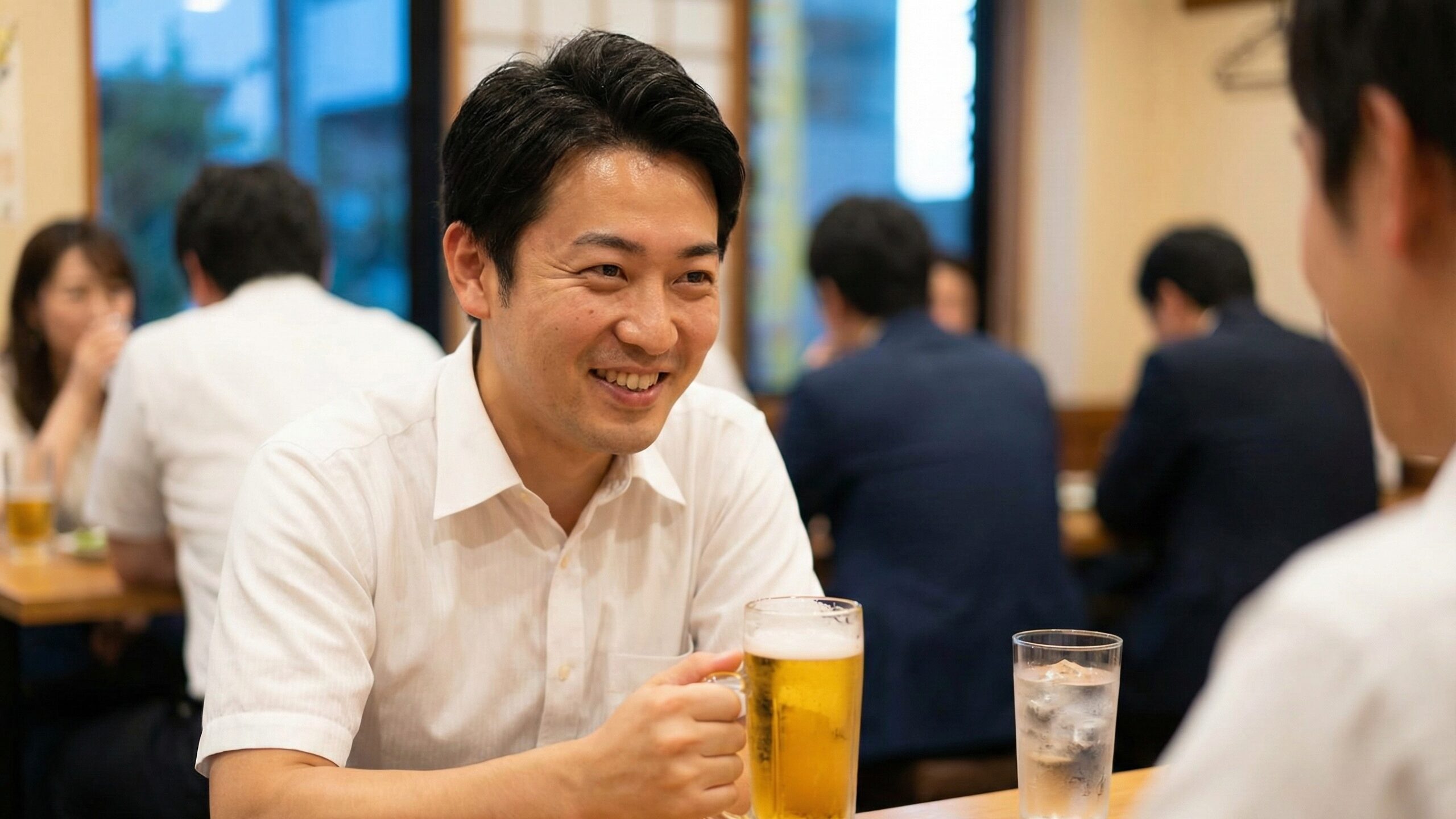 飲食店で笑顔の男性がビールのジョッキを持ち、テーブルにはチェイサーの水が置かれている様子。