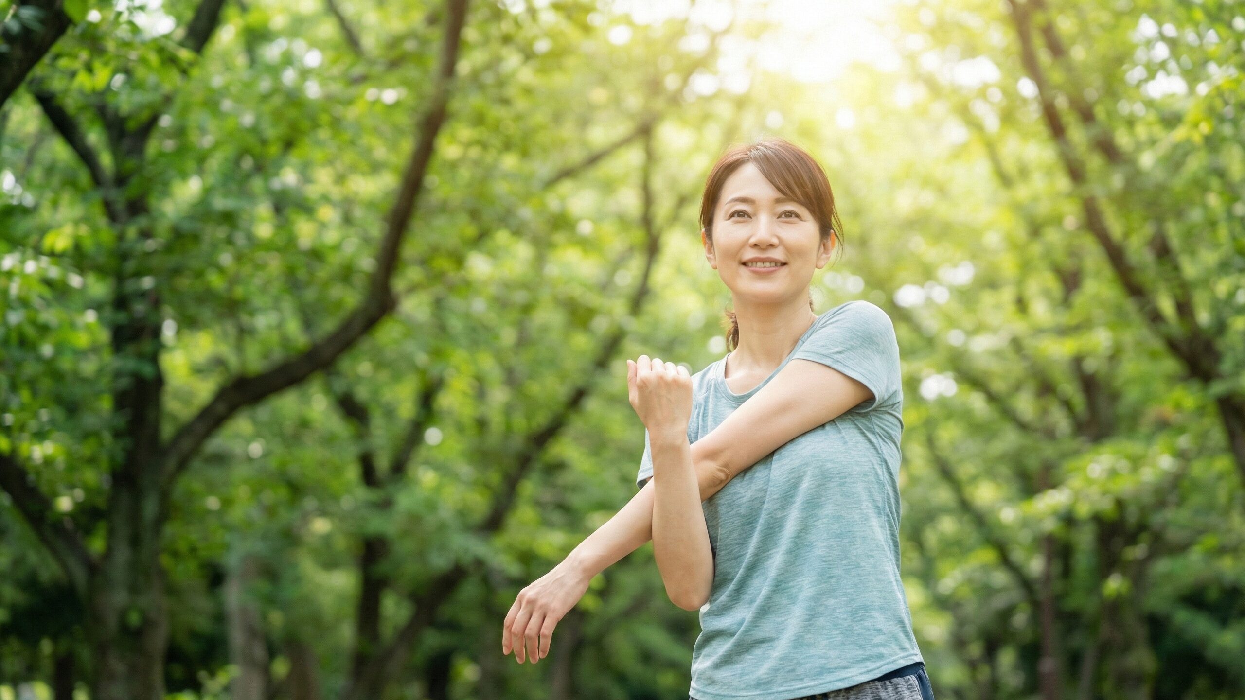 明るい日差しが差し込む新緑の公園で、腕を伸ばして心地よさそうにストレッチする女性。