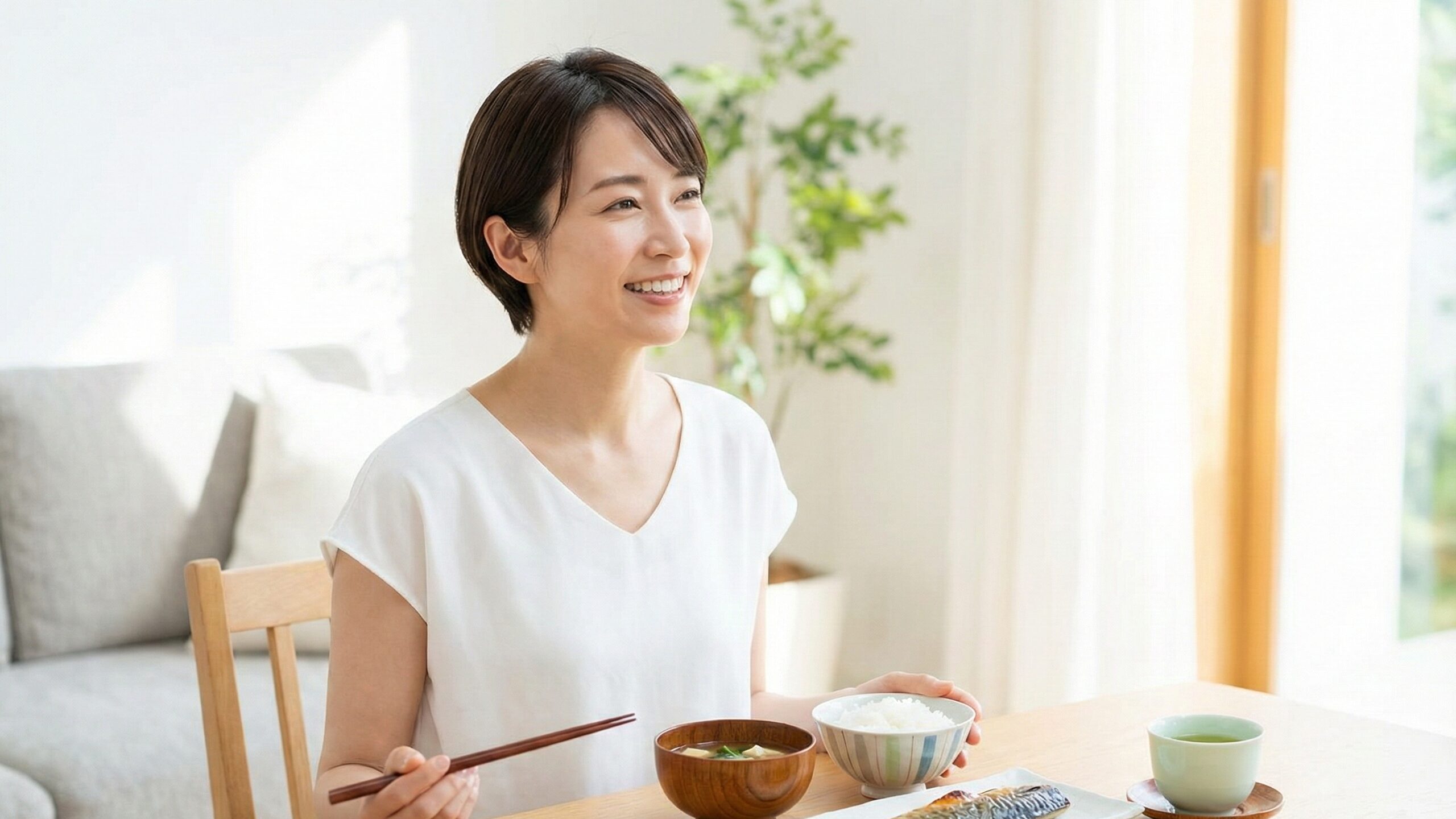 焼き魚やご飯、味噌汁が並んだテーブルを前に、箸を持って晴れやかな笑顔を見せるショートヘアの女性