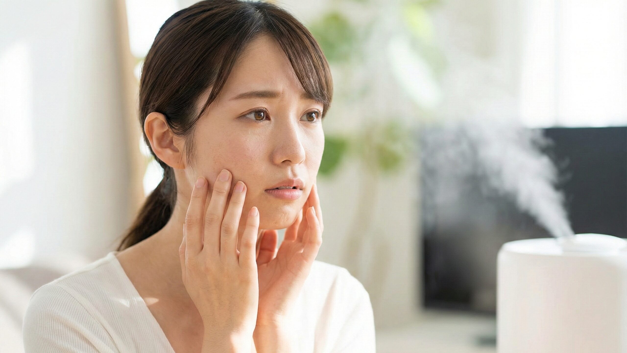 加湿器を使用しながら、カサつきが気になる頬に手を当てて鏡を見る女性