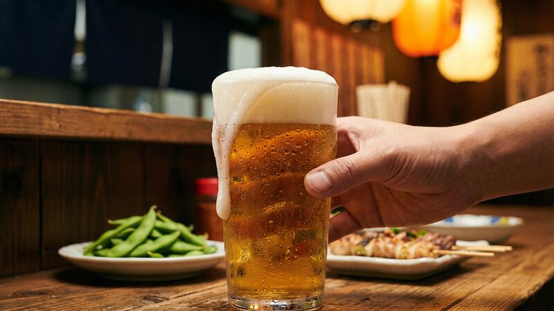 泡が溢れるビールジョッキを握る手と、皿に盛られた枝豆や焼き鳥が並ぶ賑やかなカウンター。